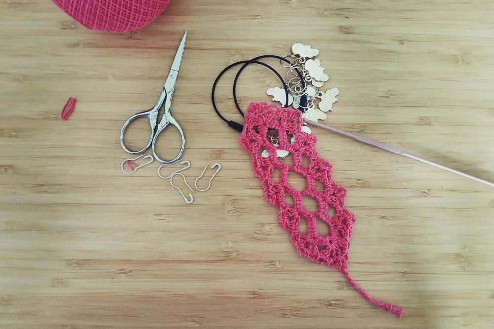 A horizontal colour photograph of a small crochet lace bookmark in a pink cotton yarn. The work is not very regular. A pink crochet hooks rests next to it as well as stitch marker with sheep attached to them. Other stitch markers are simple pins.