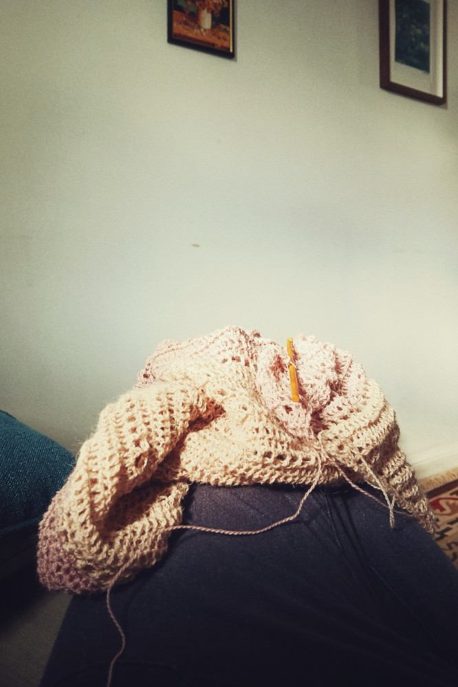 A vertical colour photograph of a crochet shawl in progress. It is made with fingering weight yarn in white, cream, and lavender so far. The work rests on my leg in a pile. I am wearing navy linen trousers. I am on a sofa barely visible in the image. The background of the image is a white wall with some frames on it.