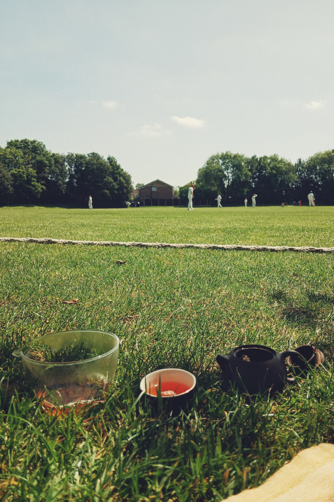 A vertical colour photograph of a gong fu tea set with teapot. The set rests on the grass at the bottom of the frame. The upper half of the frame is filled with distant figures in white playing cricket and a blue sky above.