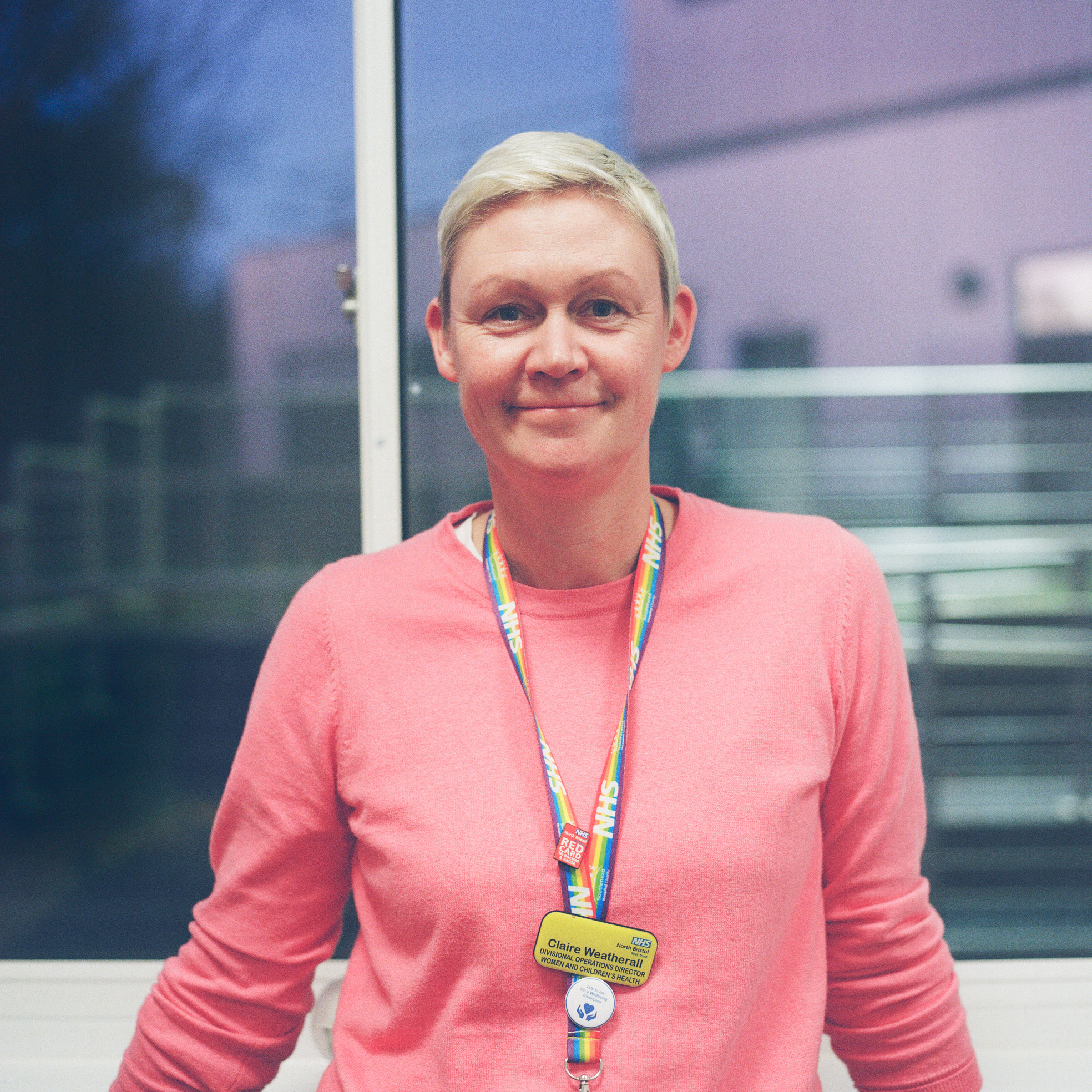 A square colour photograph of a woman from the torso up. She has short blond hair and is wearing a pink sweater. She is standing by a window in the evening.