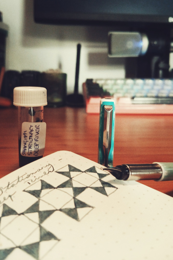 A vertical colour photograph of a fountain pen nib on a dotted notebook. Above the pen is a swatch of a grey blue ink. Above the notebook are the ink sample in a vial and the pen cap upright on a wooden desk.