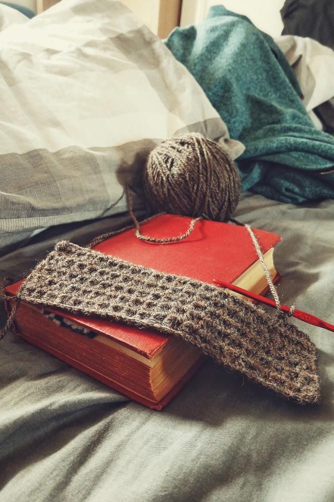 A vertical colour photograph of a brown crochet work. It's a rectangle with a red hook attached to it. The work rests on a book with an old red cover. The whole is set on an unmade messy bed.