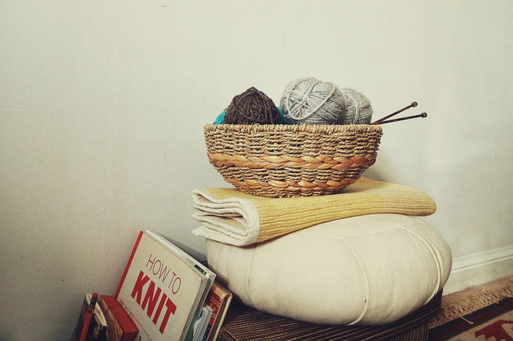 A horizontal colour photograph of a small basket with balls of yarn and a couple of wooden needles pointing out. The bowl rests on a meditation pouffe and mat on a brown box. Next to the box are some books, including one with the title 'How to Knit'.