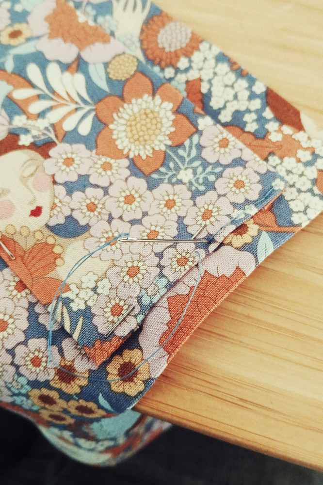 A vertical colour photograph of a needle stuck at a slight angle in fabric where stitching in a blue thread is visible. The fabric has repeated motifs of flowers and is mainly in blues and pinks and oranges.