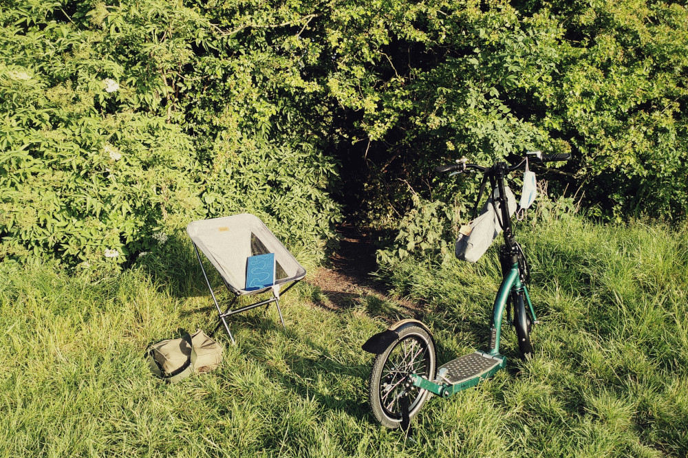 A horizontal colour photograph of a green electric scooter and a grey chair with a blue book with a wall of vegetation.