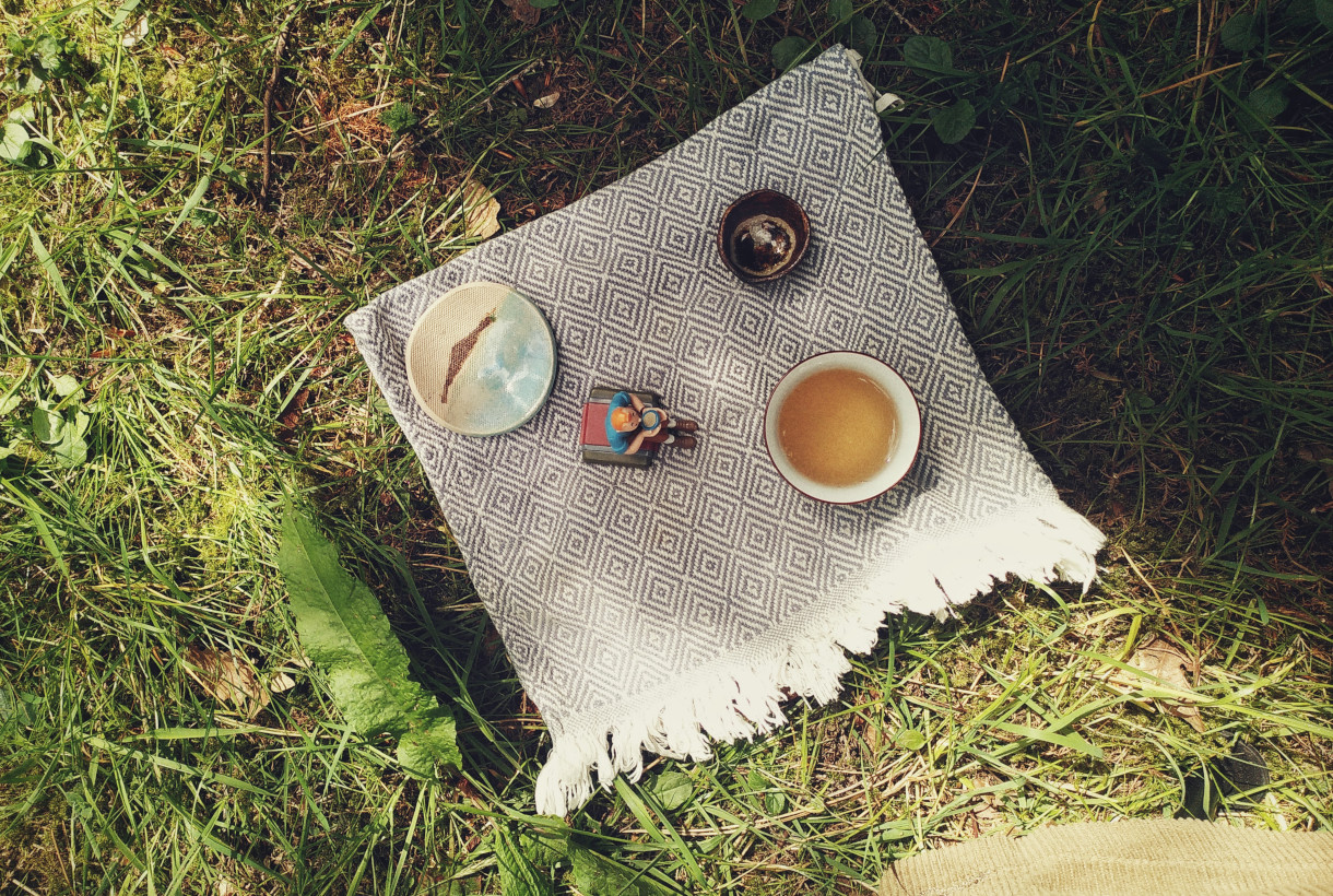 tA horizontal colour photograph of a patterned white and blue cloth on grass. On the cloth is a small cup of tea with a light coloured tea in, a couple of ceramic objects, and a Tintin figurine drinking tea.