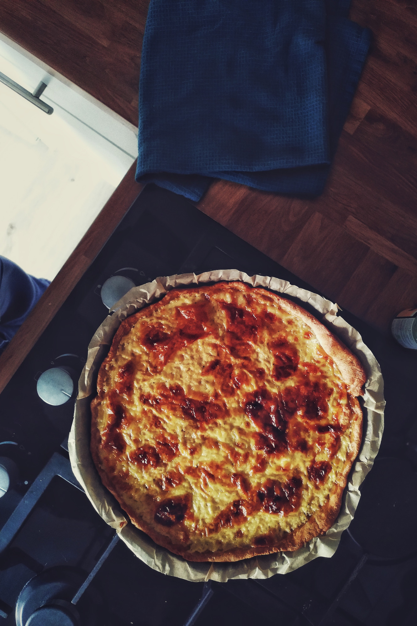 A vertical colour photograph of a cheese tart resting over a black gas hob. At the side is a hastily folded navy blue tea towel.