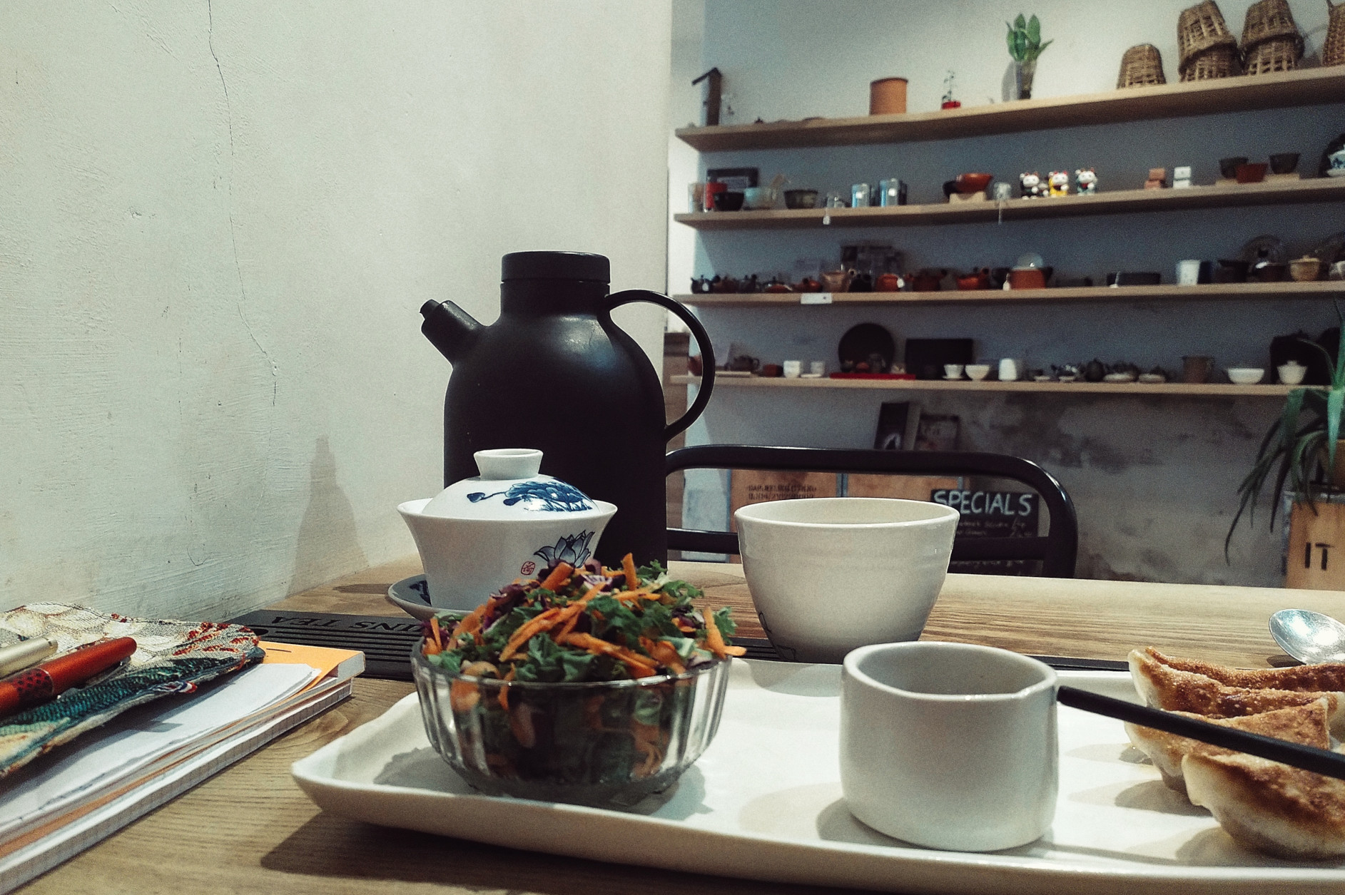 A horizontal colour photograph of a table on which rests a white tray with a small bowl of salad and some gyoza cut off by the frame, an couple of ceramic containers for tea, and some paper and pens cut off by the frame. In the background if a wall with shelves displaying traditional teaware of all sorts.