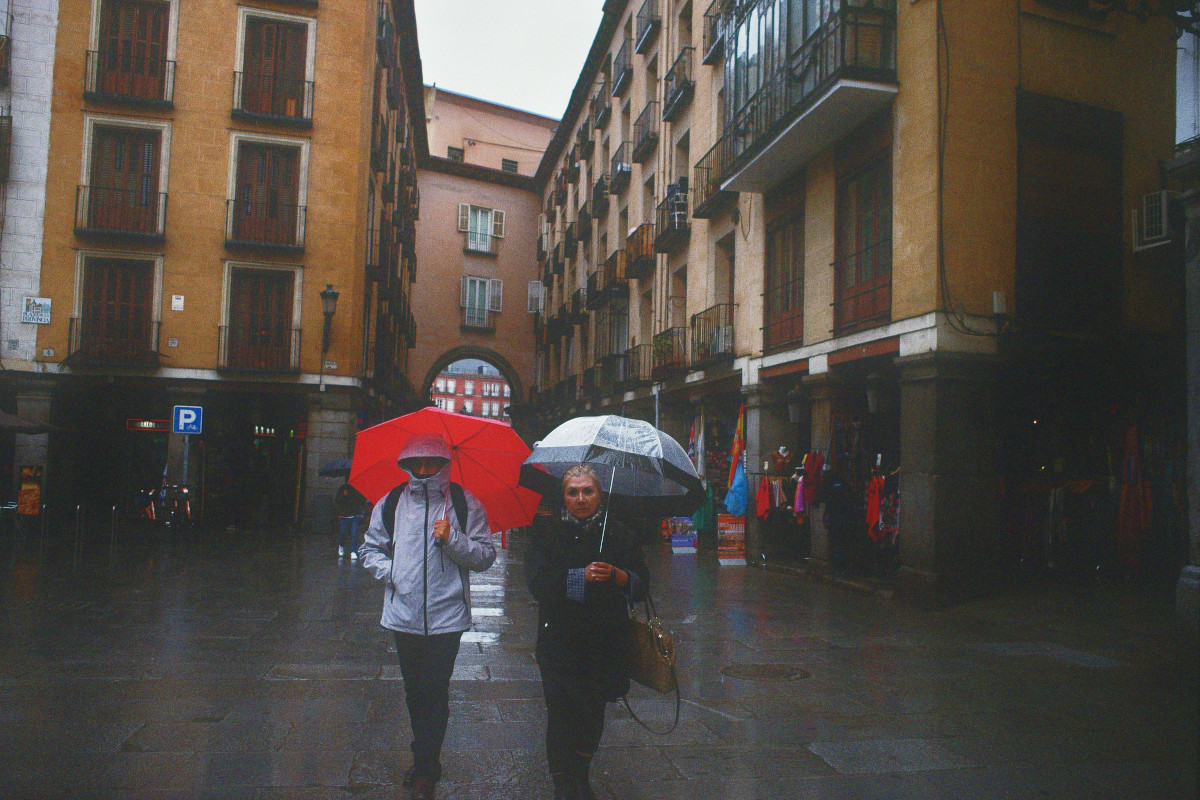 A horizontal colour photograph of two women well wrapped up and hiding under umbrellas in a street of Madrid.