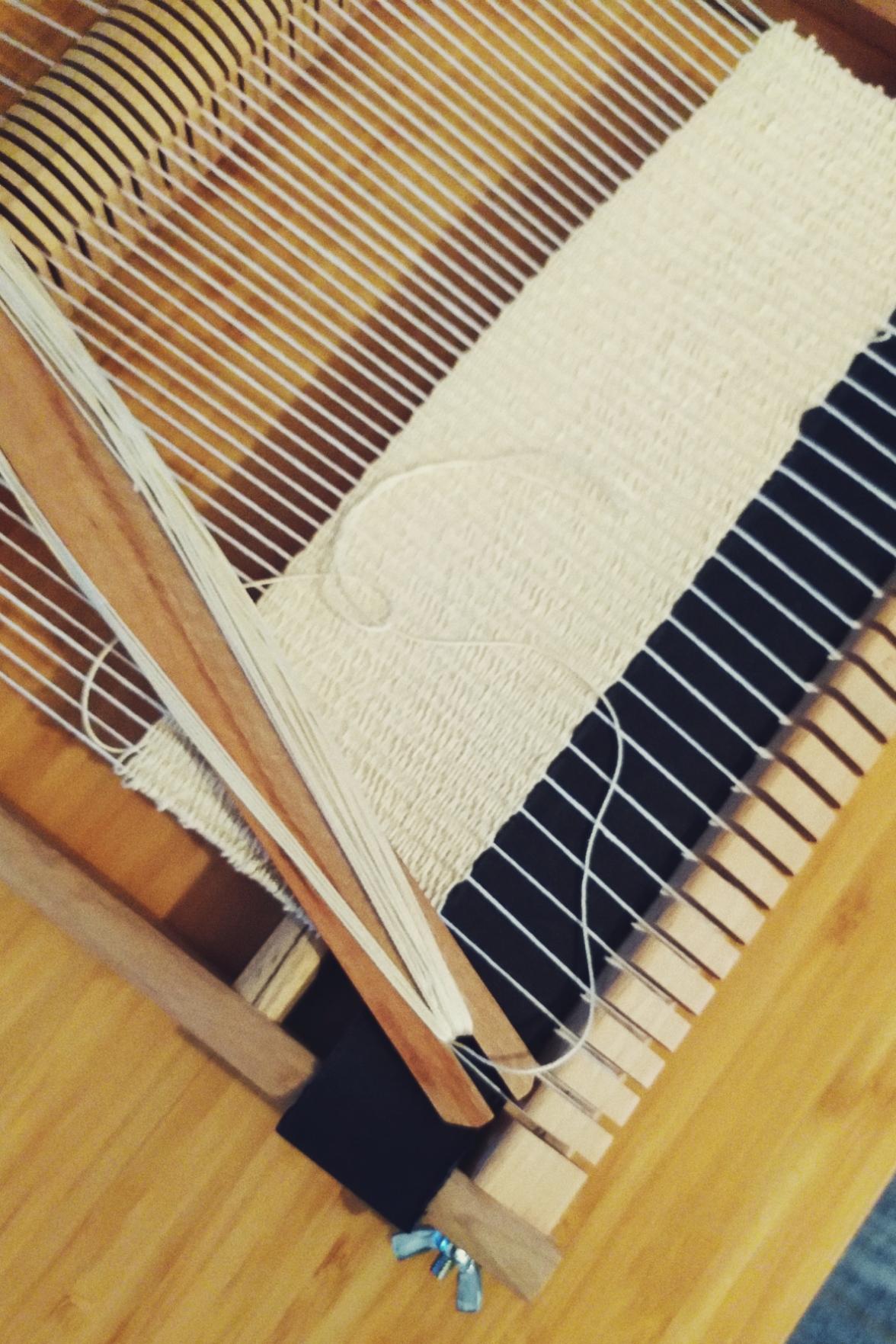 A vertical colour photograph of a wooden frame loom with a work in progress in a light creamish thin thread.