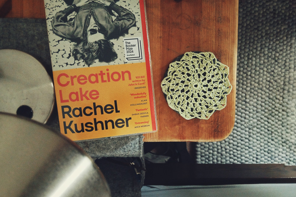 A horizontal colour photograph of a small doily in a light green thread. It rests on a dark grey coaster on the top of a bedside table. Next to it is the book 'Creation Lake' by Rachel Kushner. A metal grey lamp is visible at the bottom left corner.