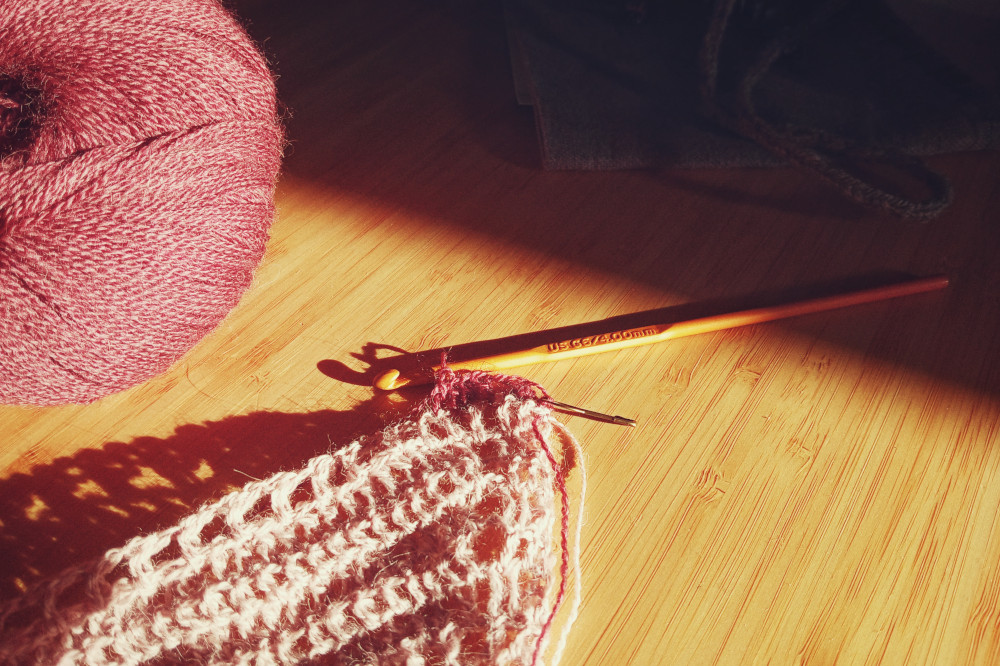 A horizontal colour photograph of the corner of a crochet work in progress. The colour has just been changed from a violet pink to a deep burgundy. The hook is orange and attached to the work. At the top left corner part of the ball of yarn is visible. A streak of light illuminate the hook and crochet with the rest of the image mostly in shadows