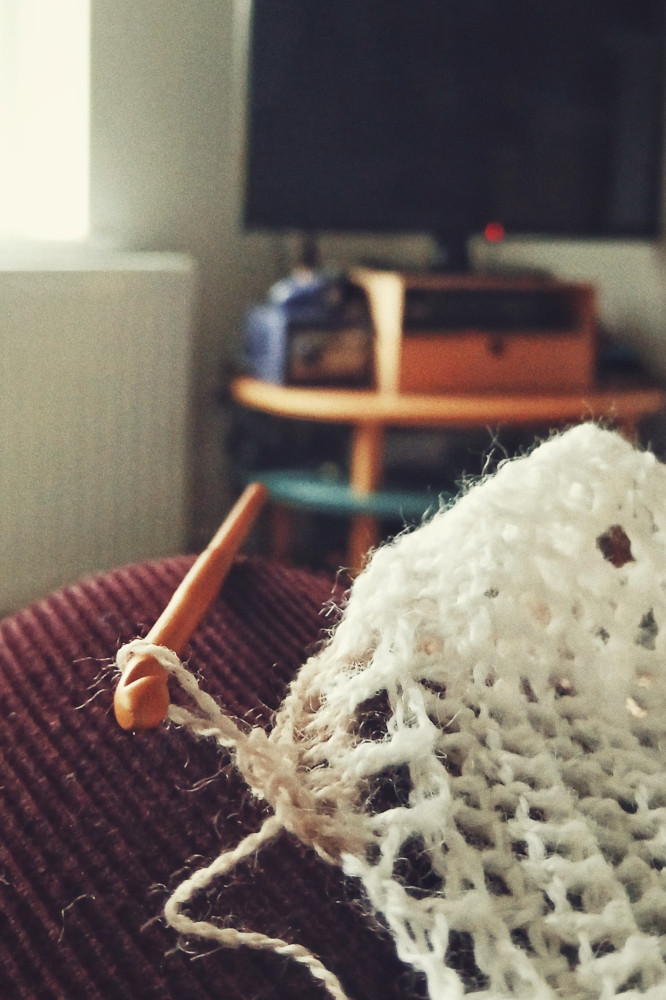 A vertical colour photograph of a crochet work. This is a close-up of a colour change with the hook holding the yarn. The old yarn in white, the new one is creamish pink colour. The work rests on my knee in burgundy corduroy. Behind, blurred is a living room with a TV turned off.