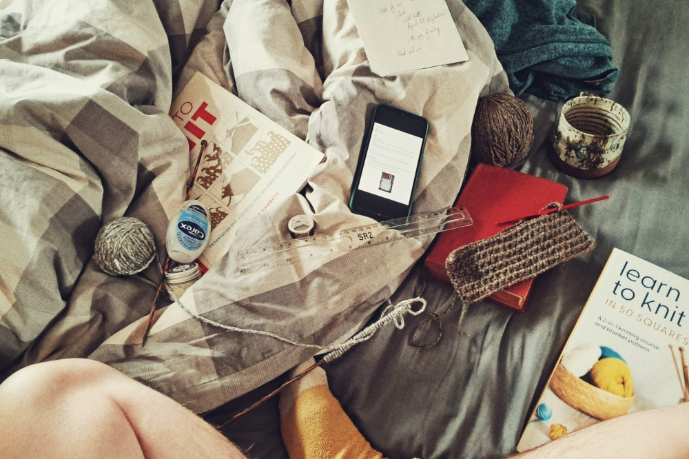 A horizontal colour photograph of a messy bed with books about knitting, needles with the start of a grey work on, a brown crochet work in progress on a book by a mug of tea. My crossed legs in yellow and white socks are poking at the bottom of the frame.