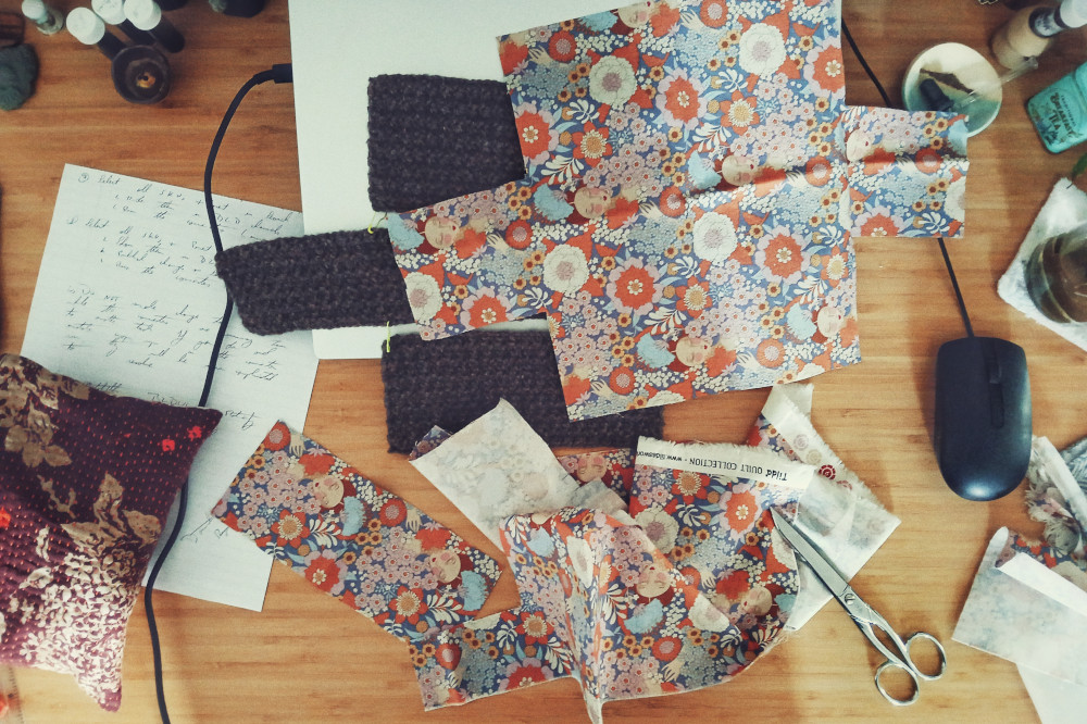 A horizontal colour photograph of a messy desk with fabric cut to size, scissors, crochet work, laptop, and other desk paraphernalia all over the place. The focus is the fabric work. The fabric is all blues and pinks and oranges with repeating pattern of leaves and a woman's face.