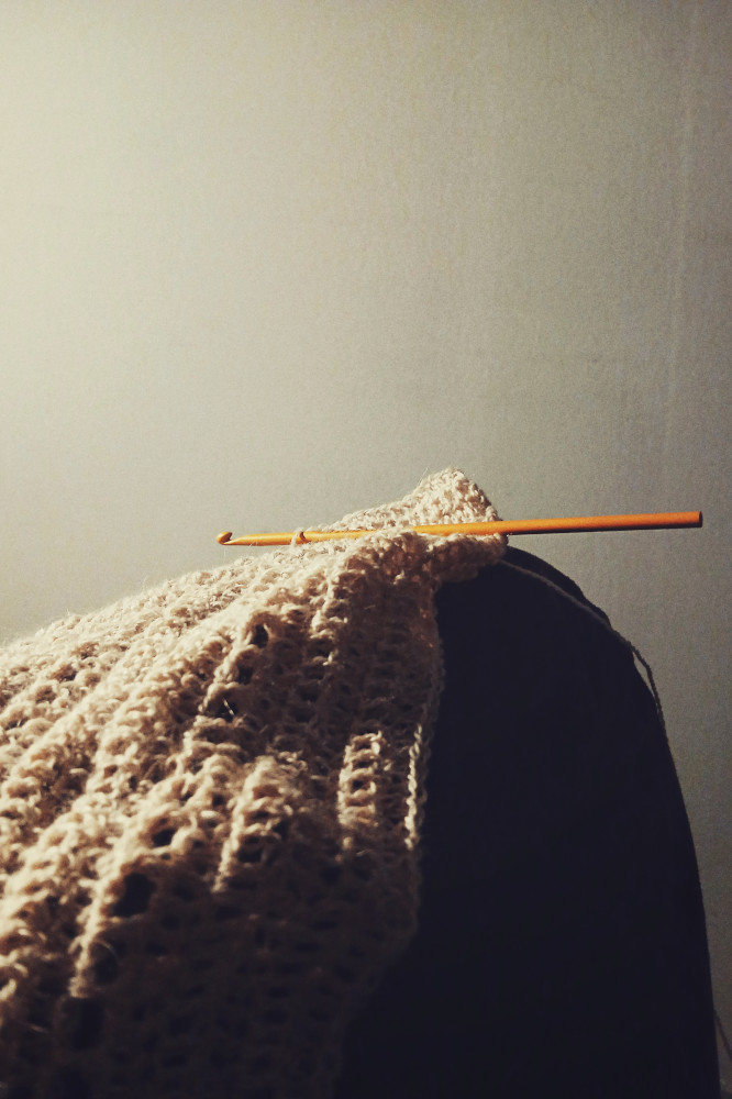 A vertical colour photograph of a crochet shawl in progress on my knee. The crochet hook is orange and rests perfectly still at the highest point of my knee.