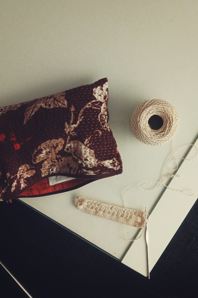 A vertical colour photograph of a filet crochet work in progress on a white table. The hook is on the work. The thread ball is above it. Next to the thread and work is a red pouch with a flower pattern and a running stich across the entire surface.