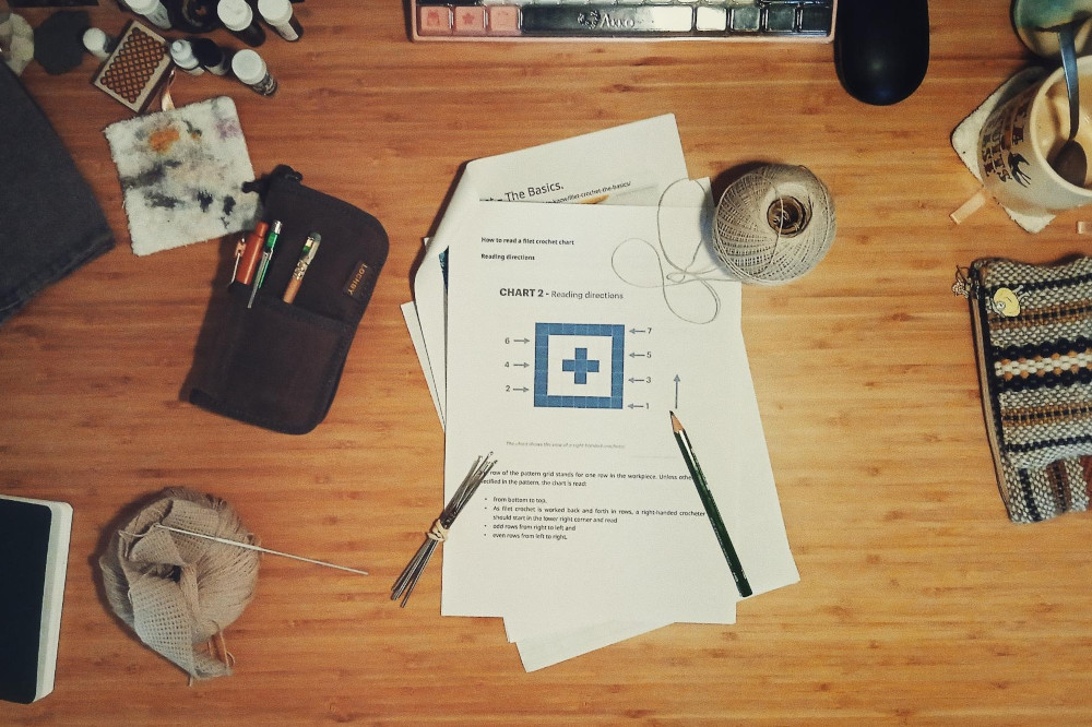 A horizontal colour photograph of a desk with a stack of printed paper. The op one shows a filet crochet chart. At the corners of the paper is a ball of cotton thread, some fine steel crochet hooks, and a pencil. Also on the desk is a pen pouch, another crochet work u progress tucked in a creamish ball of thin wool yarn, an empty jug, ink samples, etc.