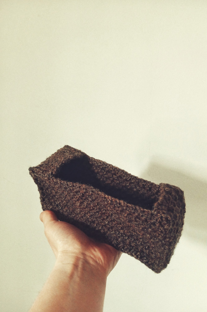 A vertical colour photograph of my white hand holding up a rectangular crochet pouch in front of a white wall.