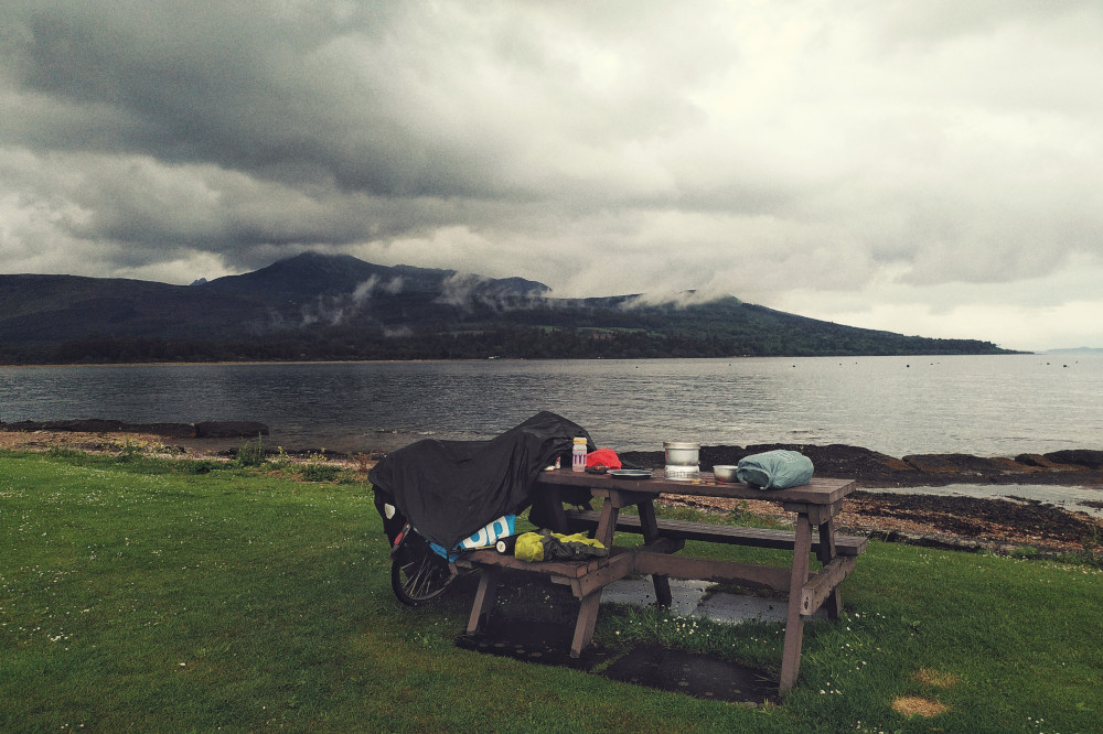 A horizontal colour photograph of a strip of grass by the sea. In the distance are hills covered by clouds. On the grass is a picnic table with a camping stove and other camping kitchen paraphernalia. At the side of the table a bicycle is resting under a dark green canva poncho.