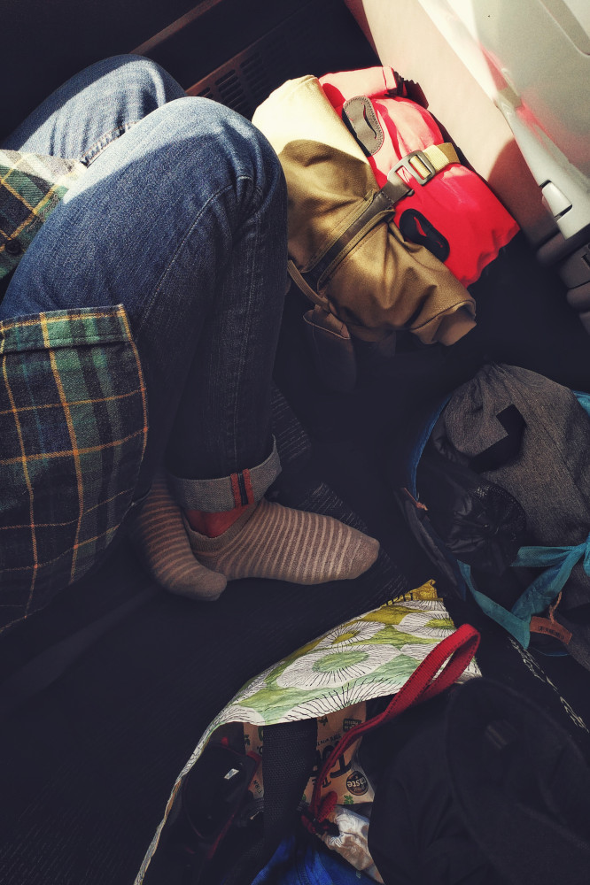 A vertical colour photograph of my legs in blue jeans curled on a train seat. Around me, on the floor and the seat next to me, are bags filled with camping gear.