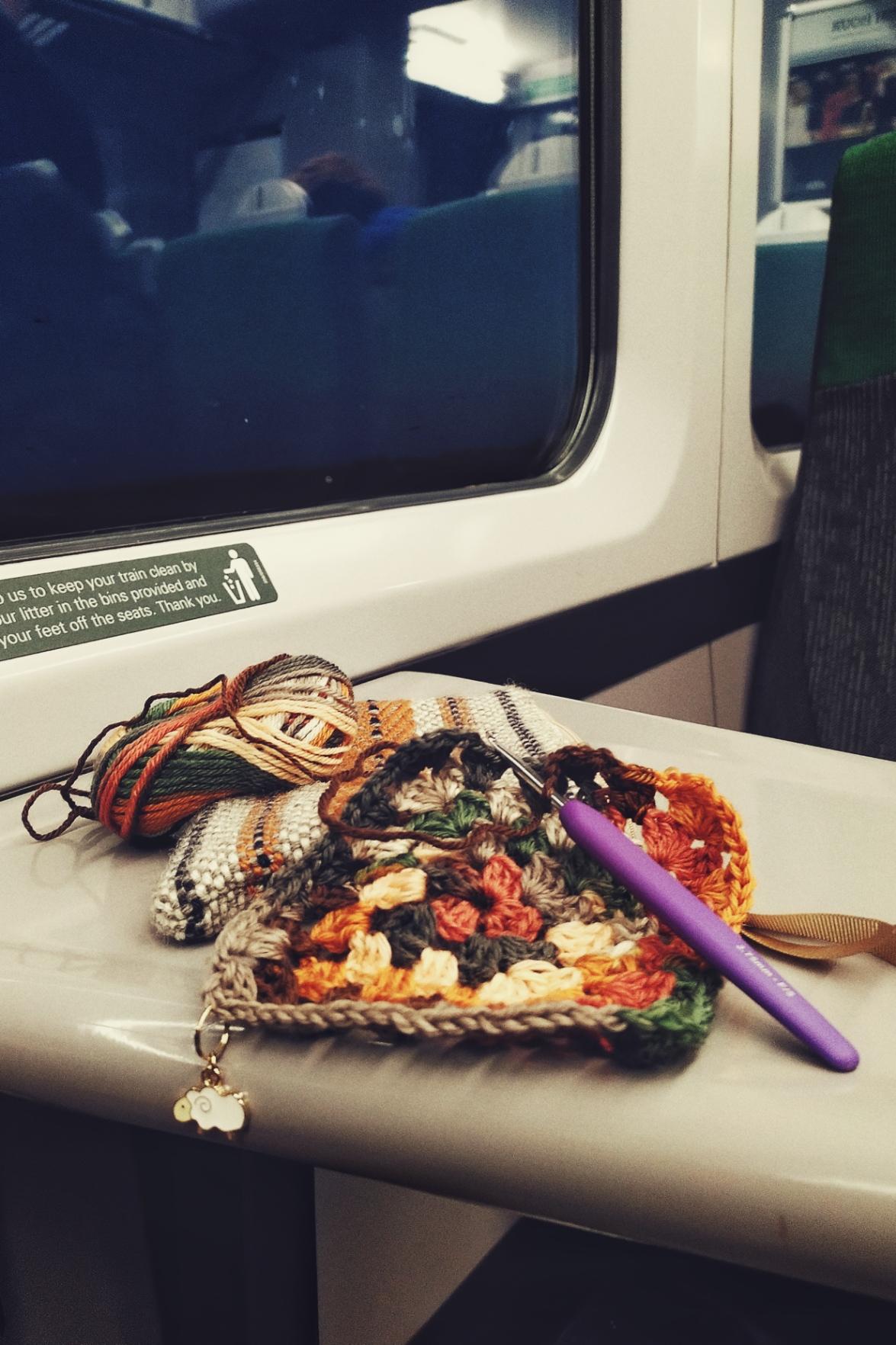 A vertical colour photograph of a multicoloured granny square in progress on a small white train table. A purple crochet is attached to the square. The multicoloured yarn rests on a cream, orange, and black woven bag.