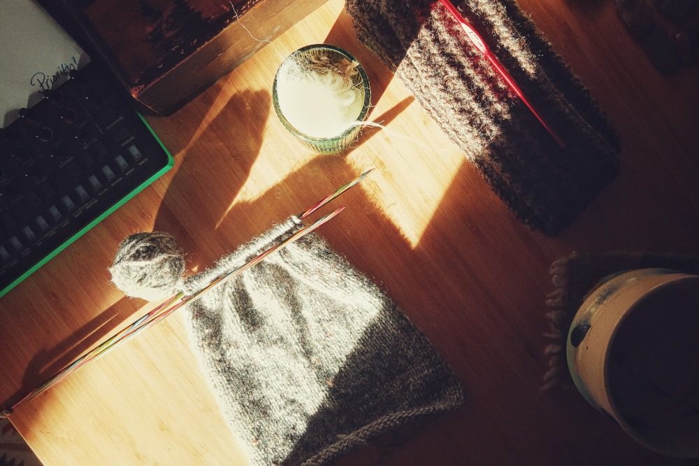 A horizontal colour photograph of a small grey knitted rectangles on wooden needles. Next to them is a small glass pot full of discarded wool. The wool is white and overblown in the image. Further back is a brown crochet rectangle with a red crochet on it.