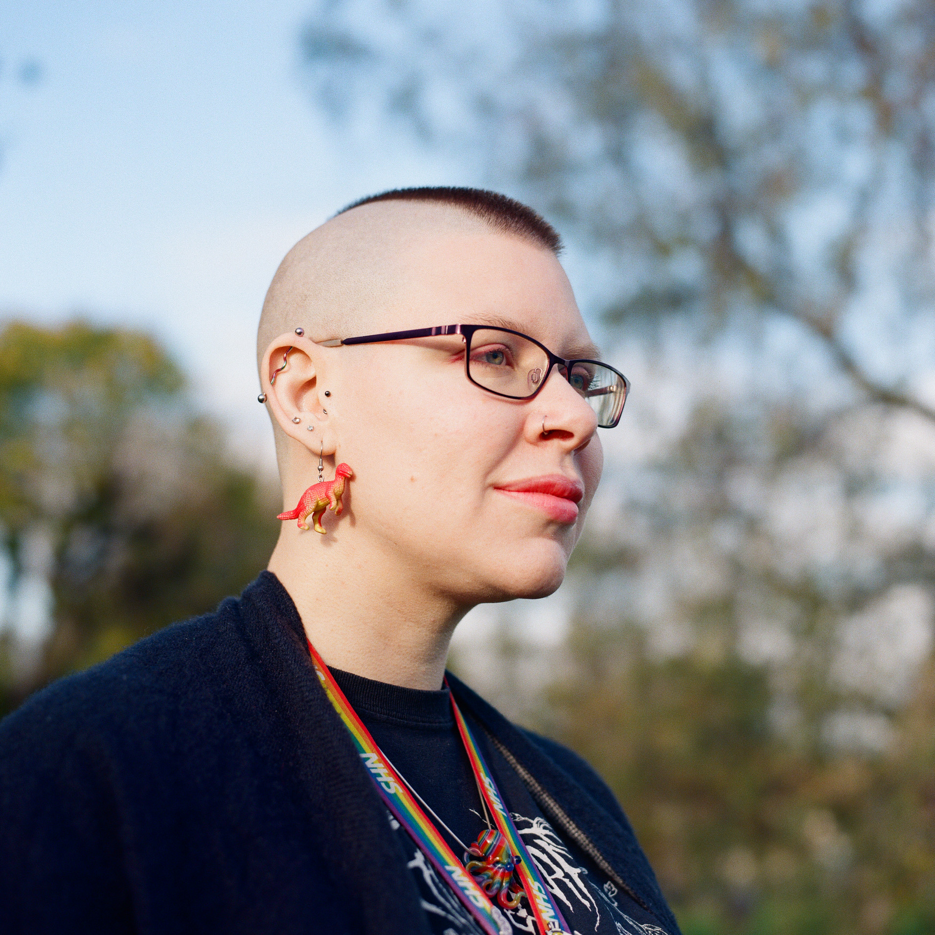 A colour photograph of a person from the shoulders up. They are have short cropped hair and dinosaur earrings. They are looking off in the distance.