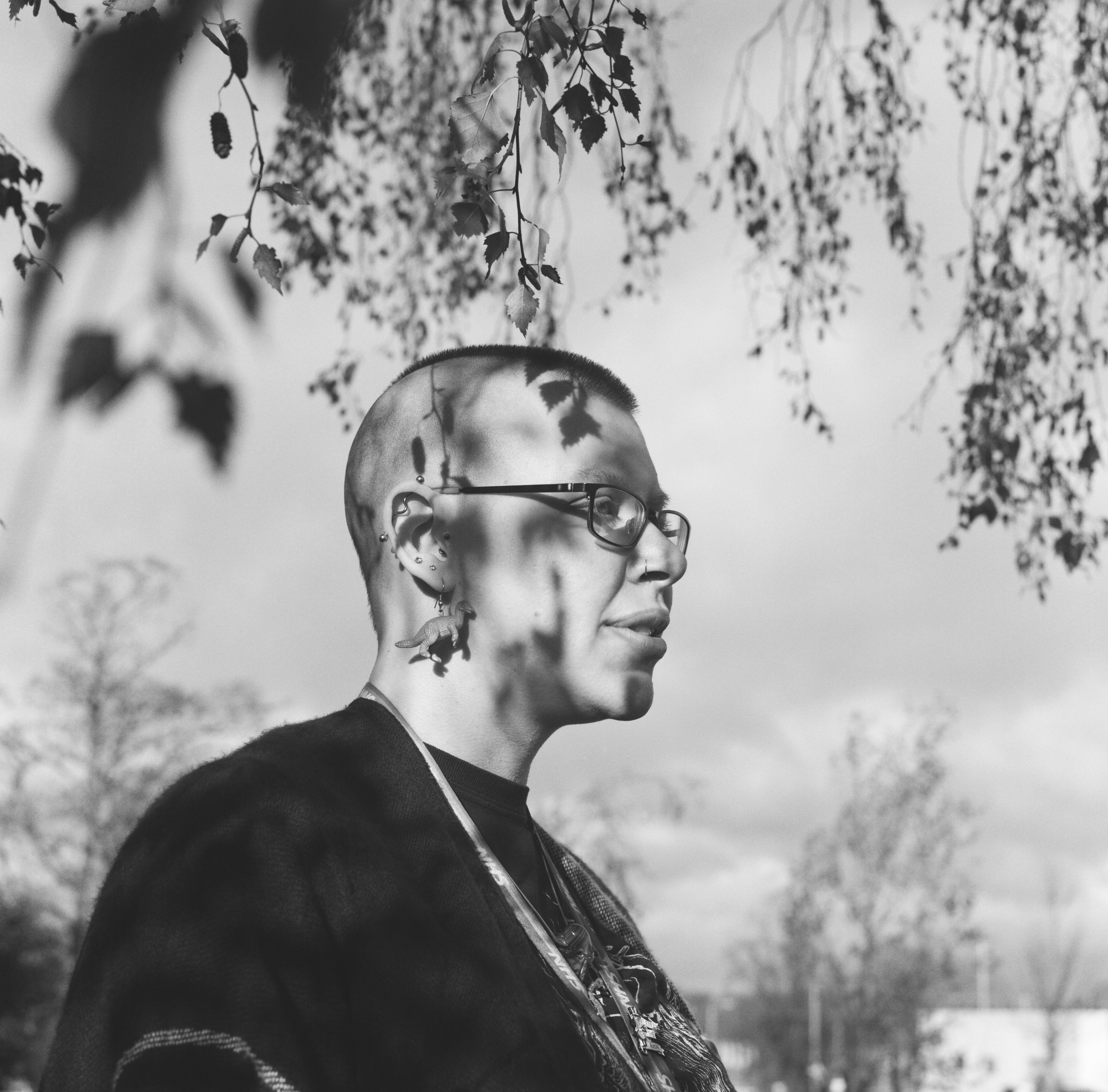 A black and white photograph of a person from the shoulders up. They are standing in the midst of branches. They have short cropped hair. Shadows of the leaves fall cross their faces.