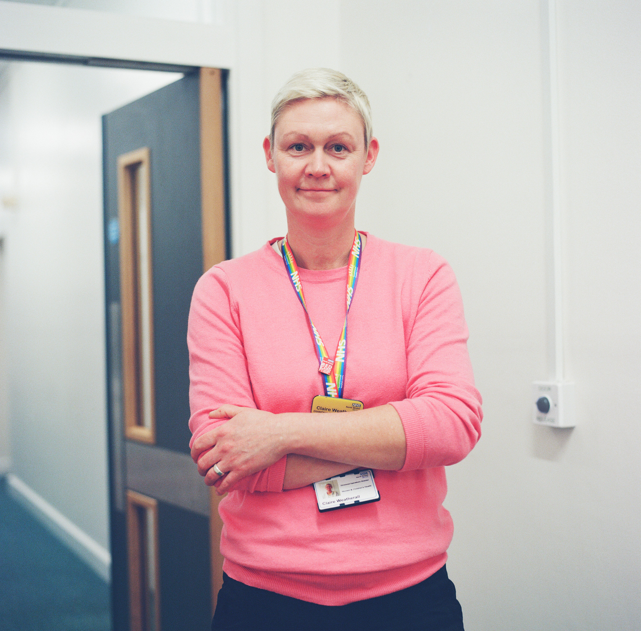 A square colour photograph of a woman from the waist up. She has short blond hair and is wearing a pink sweater. She is standing arms folded in a corridor.