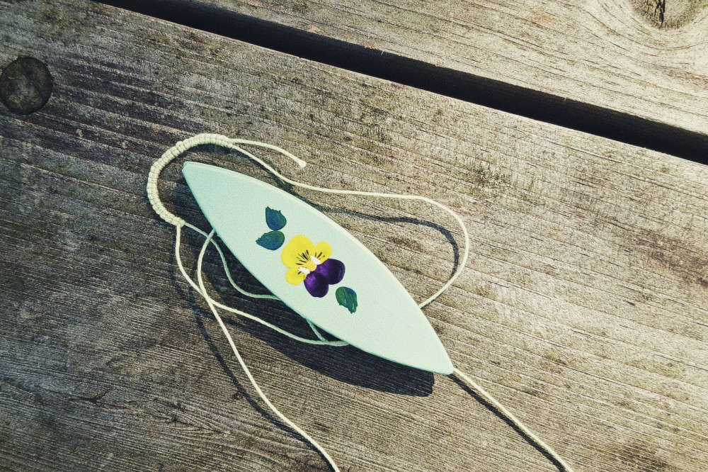 A horizontal colour photograph of a green tatting shuttle with a pansy painted on it. It rests on a wooden table. By the shuttle is a row of tatted knots in a light green colour.