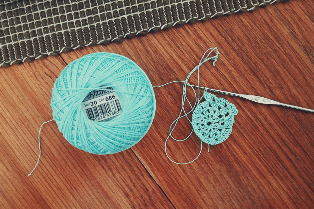 A horizontal colour photograph of a small doily made with a steel crochet hook rest on a wooden table. The doily is only just started. The ball of thread is a turquoise colour and is next to the doily. Above the work and thread is a woven a tiny bit of a mat in dark green.