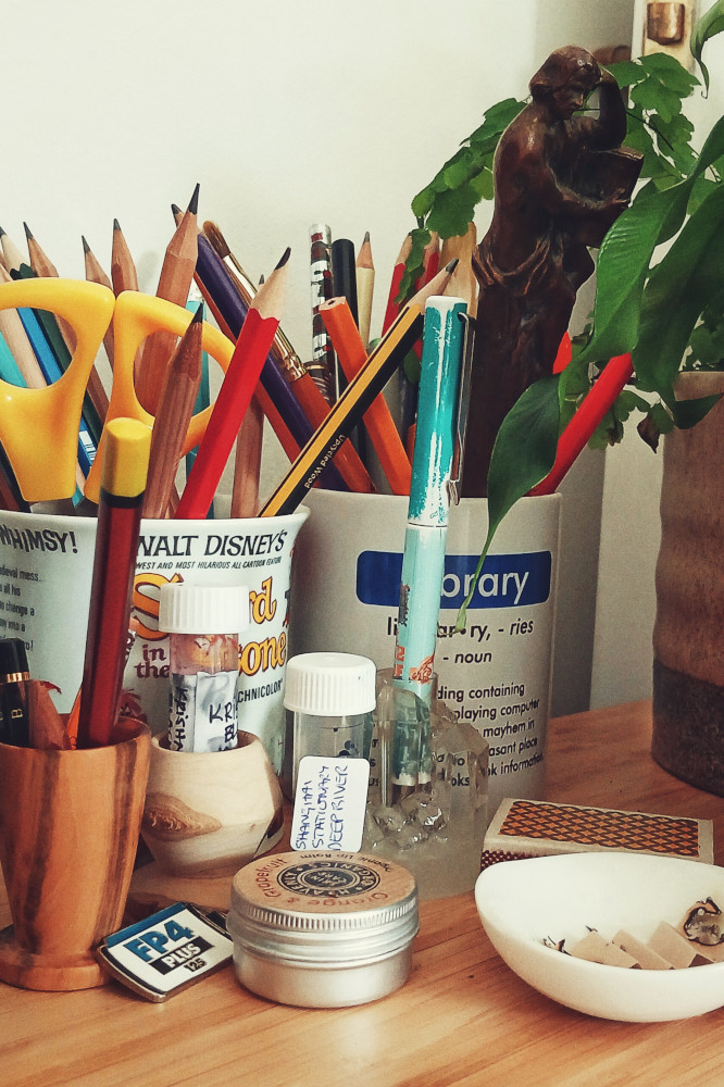 A vertical colour photograph of a teal and light green fountain pen in a translucent pen holder. The pen by two mugs holding wooden pencils. Around are small wooden turned holder, a film photography pin, and a round small tin of lip balm.