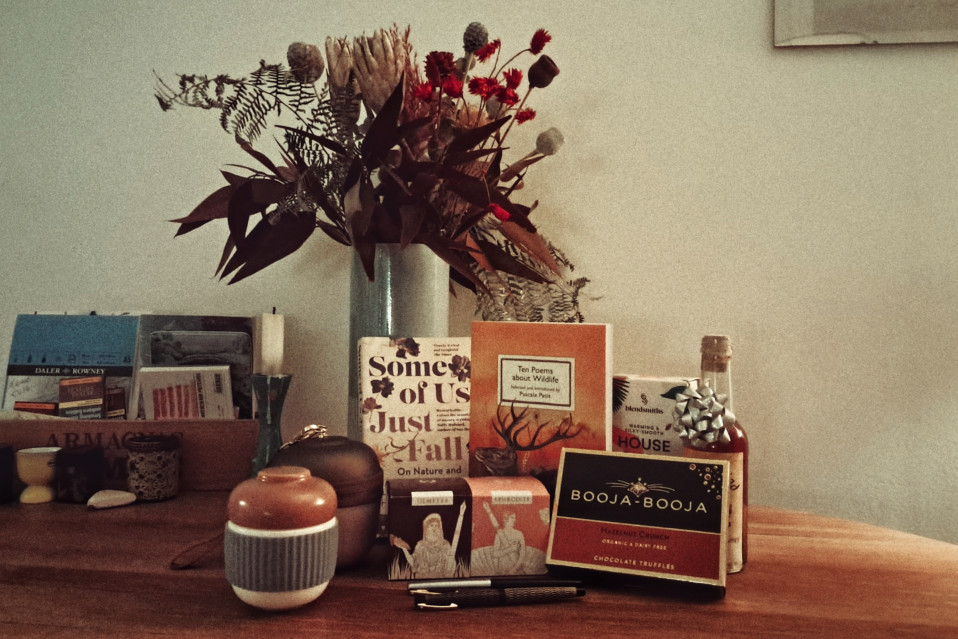 A horizontal colour photograph of a object presented on a wooden table. There are books, fountain pen inks, pens, a travel tea pot, a bottle of mead, some chocolate. In the background a grey vase holds an assortment of reddish dried flowers.