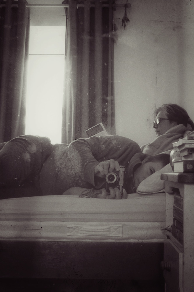A vertical sepia photograph of me (a white person with short dark hair in a zipped hoodie and dark linin trousers) lying in bed. I'm holding a camera towards the mirror to create this image. My eye is half closed. Behind me is a white woman with dark hair and glasses. She is sitting up, wears a dressing gown, and is reading a book. Behind are a white wall and open curtains that let in a glowing light. Just visible on the right hand corner is a bedside table with books all over.