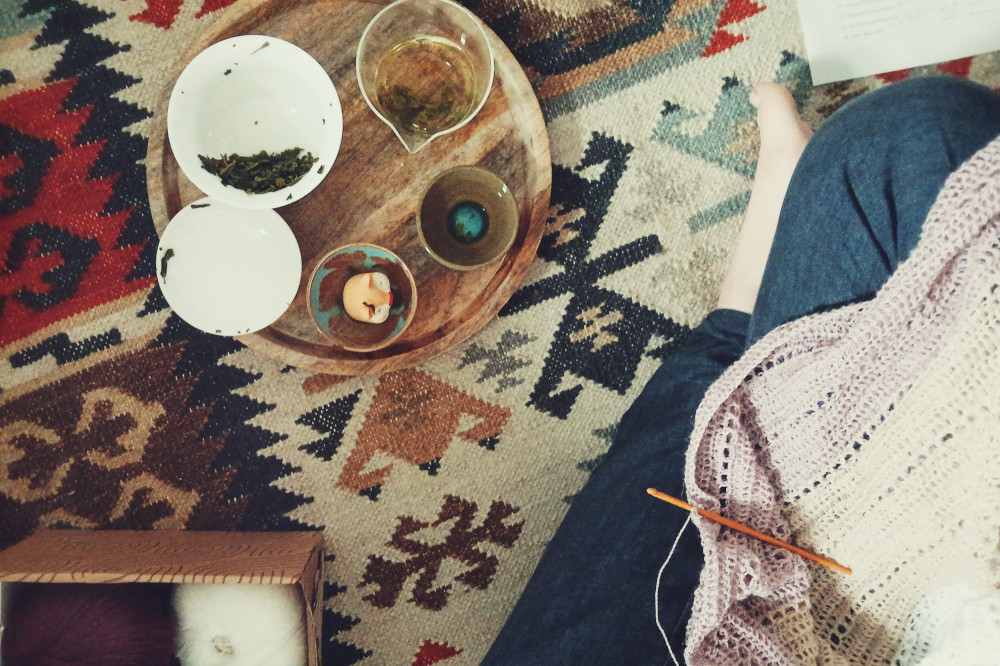 A horizontal colour photograph split in two halves. On the left hand side towards the top is a round wooden tray with gong fu tea equipment on it. On the right hand side towards the bottom are my crossed legs in a navy linen trouser and bare feet. On my legs is part of a crochet work in progress. The scene is on the floor, over a colourful carpet with geometrical shapes.