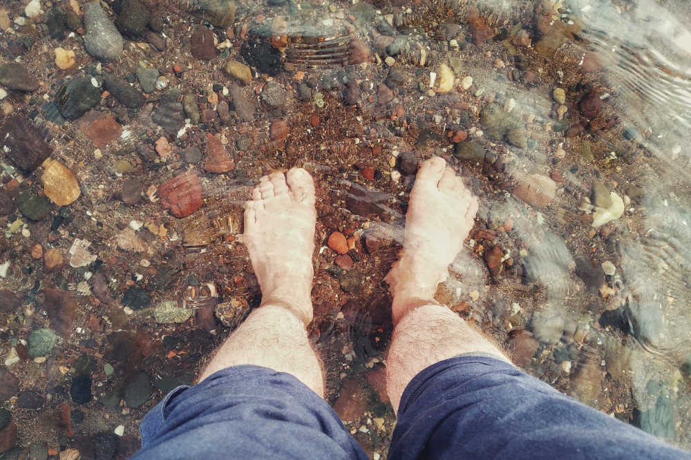 A horizontal colour photograph of my feet in calm transparent water. The ground is made out of pebbles of various size and colours. My leg are bare and hairy, my dark blue linen trousers rolled up.
