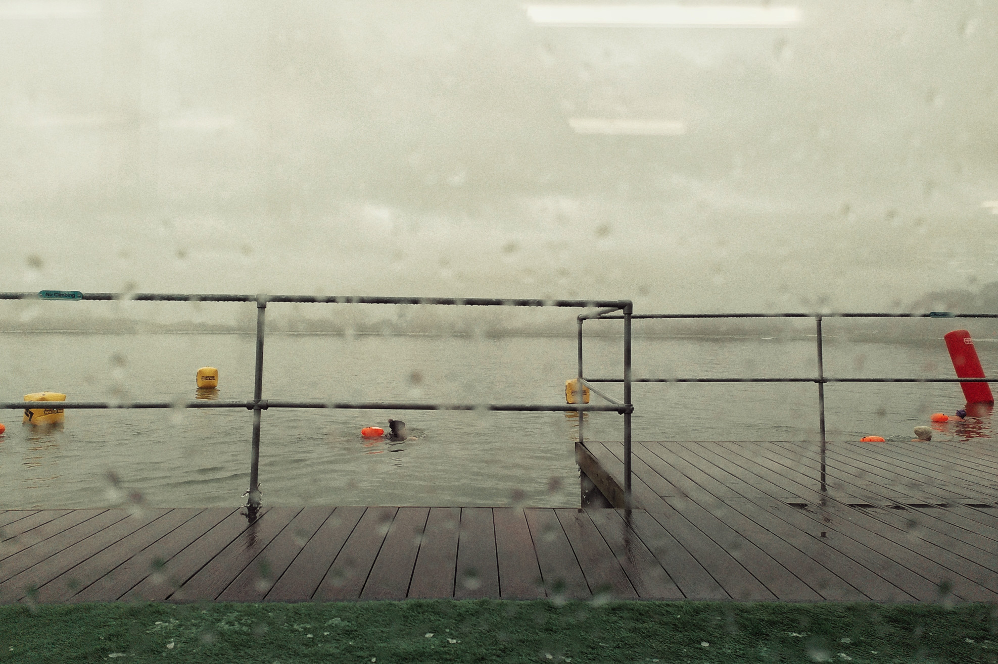 A horizontal colour photograph of a lake with people swimming in it seen through a window splattered with rain drops.