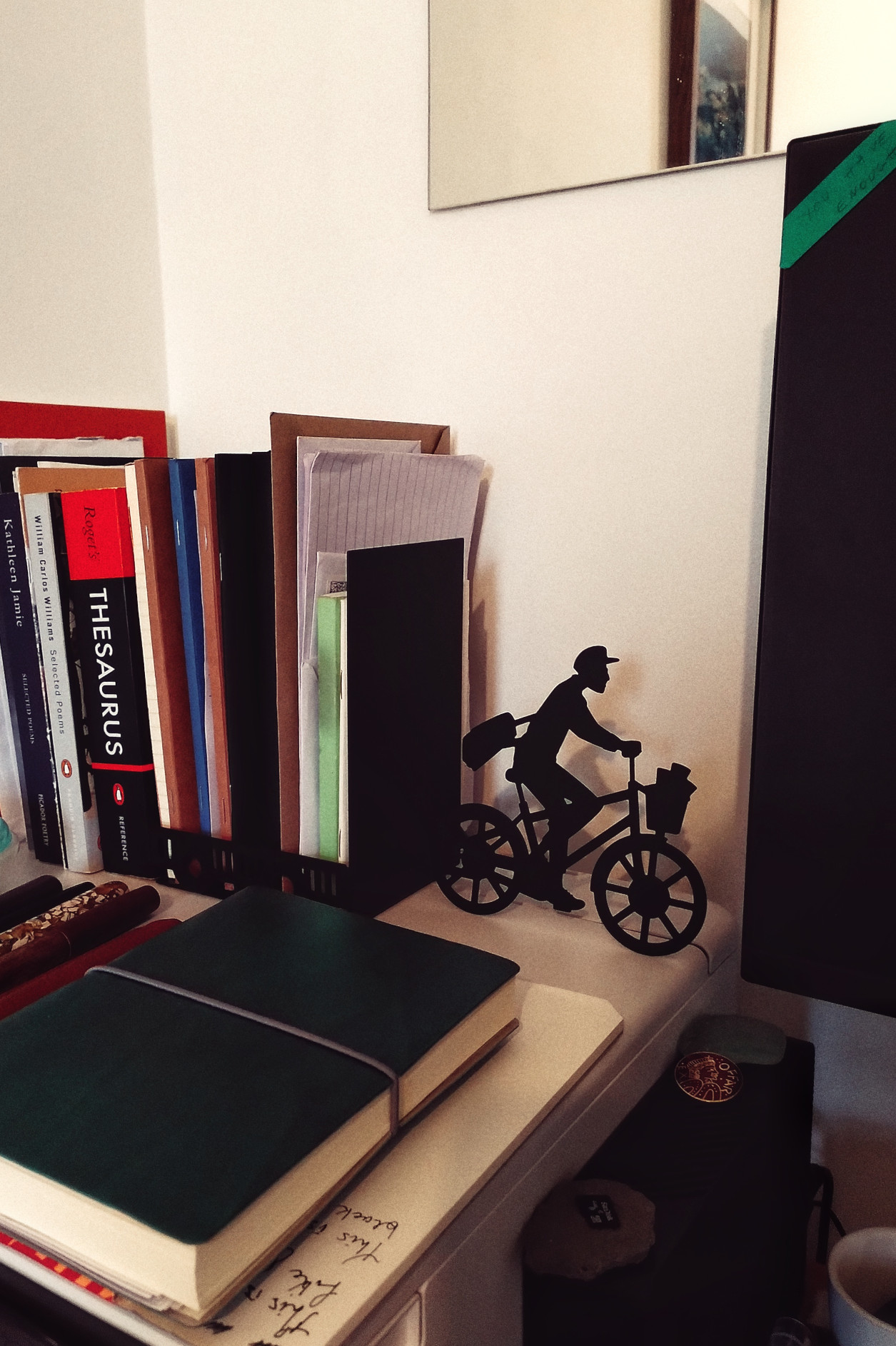 A room corner with a pile of books on a printer held by a bookend. The bookend is black and spearheaded by the figure of a cyclist. On the right edge of the image is the side of a black computer screen. In the foreground is a small pile of paper topped with a notebook.