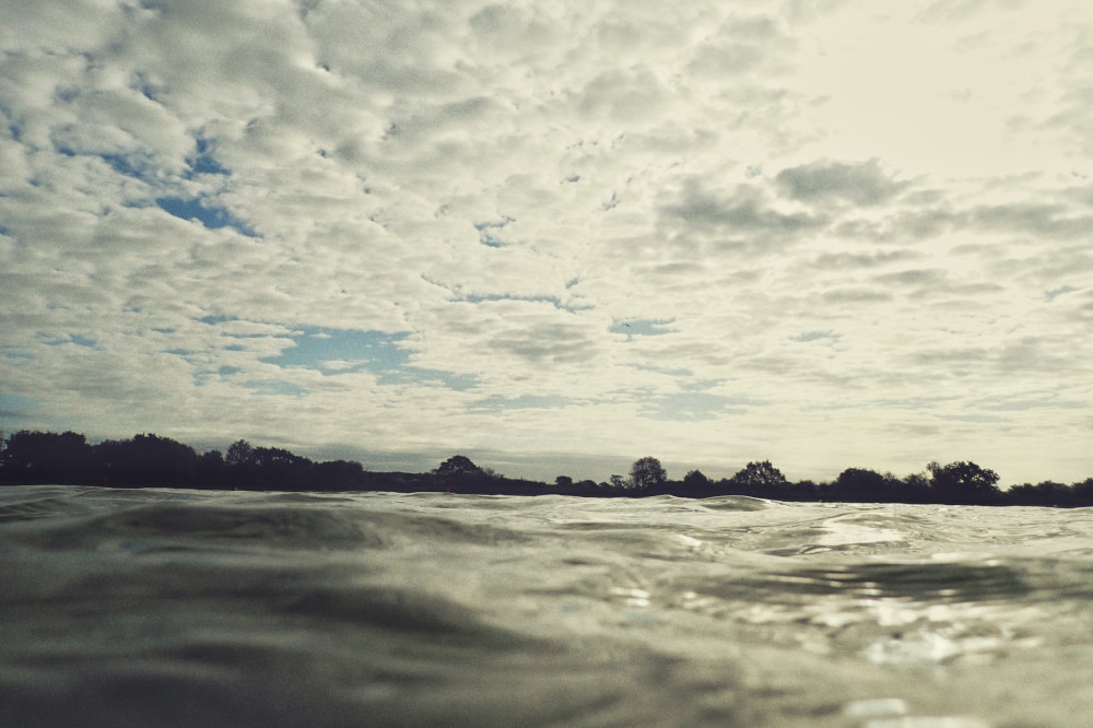 A horizontal colour photograph of the water of a lake as seen from the water. In the distance is a line of trees. The sky is a patchwork of clouds.