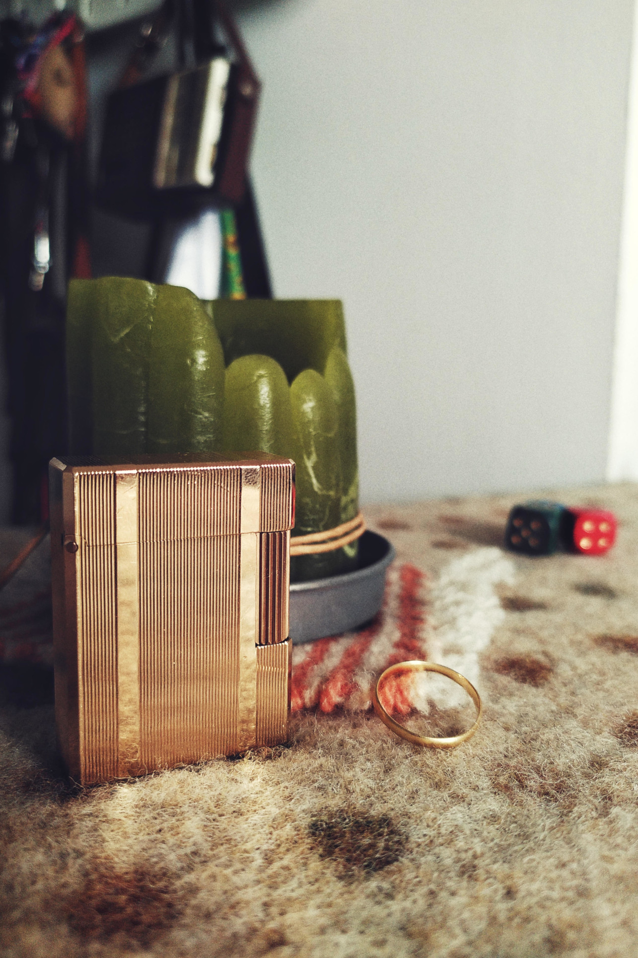 A vertical colour photograph of a gold plated rectangular lighter by a half used thick round green candle. The candle rests on a pink and white woolen coaster. Next to those is a thin golden wedding band.