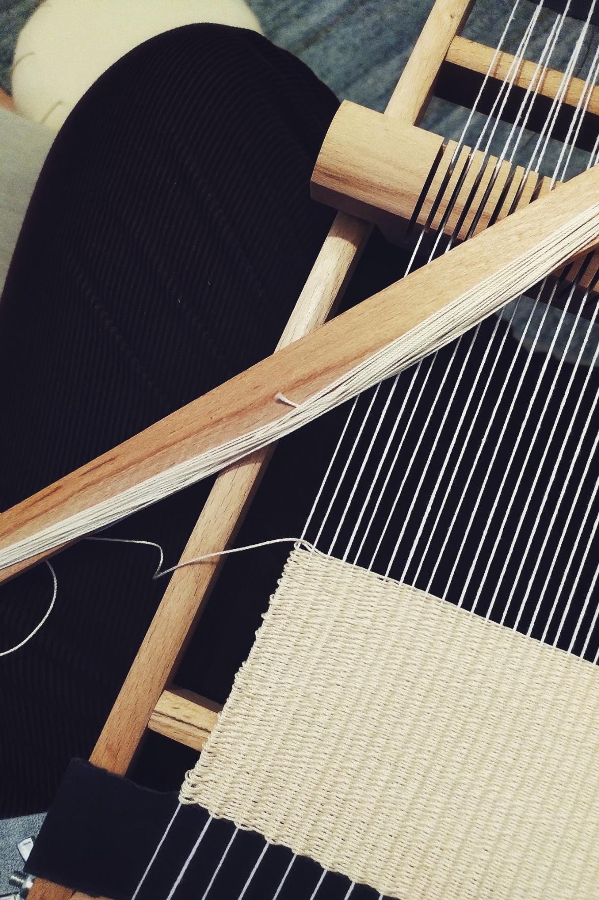 A vertical colour photograph of part of a wooden frame loom resting on my lap. I am wearing dark burgundy corduroys. The weave on the frame in with a cream thin thread. The shuttle rests diagonally across the frame.