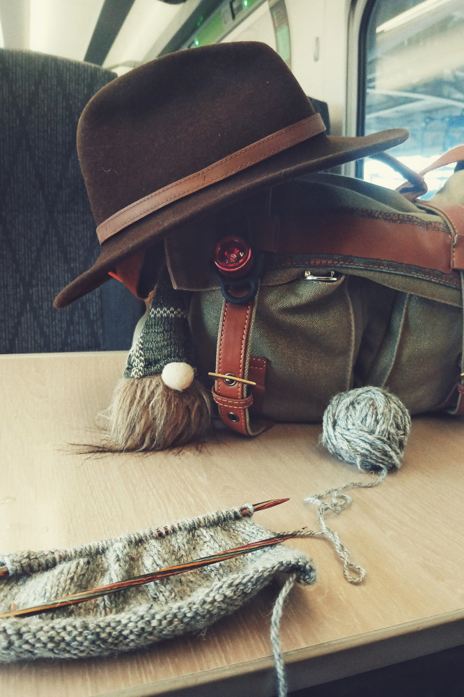 A vertical colour photograph of a train table by dark grey seats. On the table is a green bag with a dark brown felt hat resting on it. Attached to the bag is the head of a gnome. In front of the bag is a knitting work in progress with a grey yarn. The ball rests between the needled and the bag.