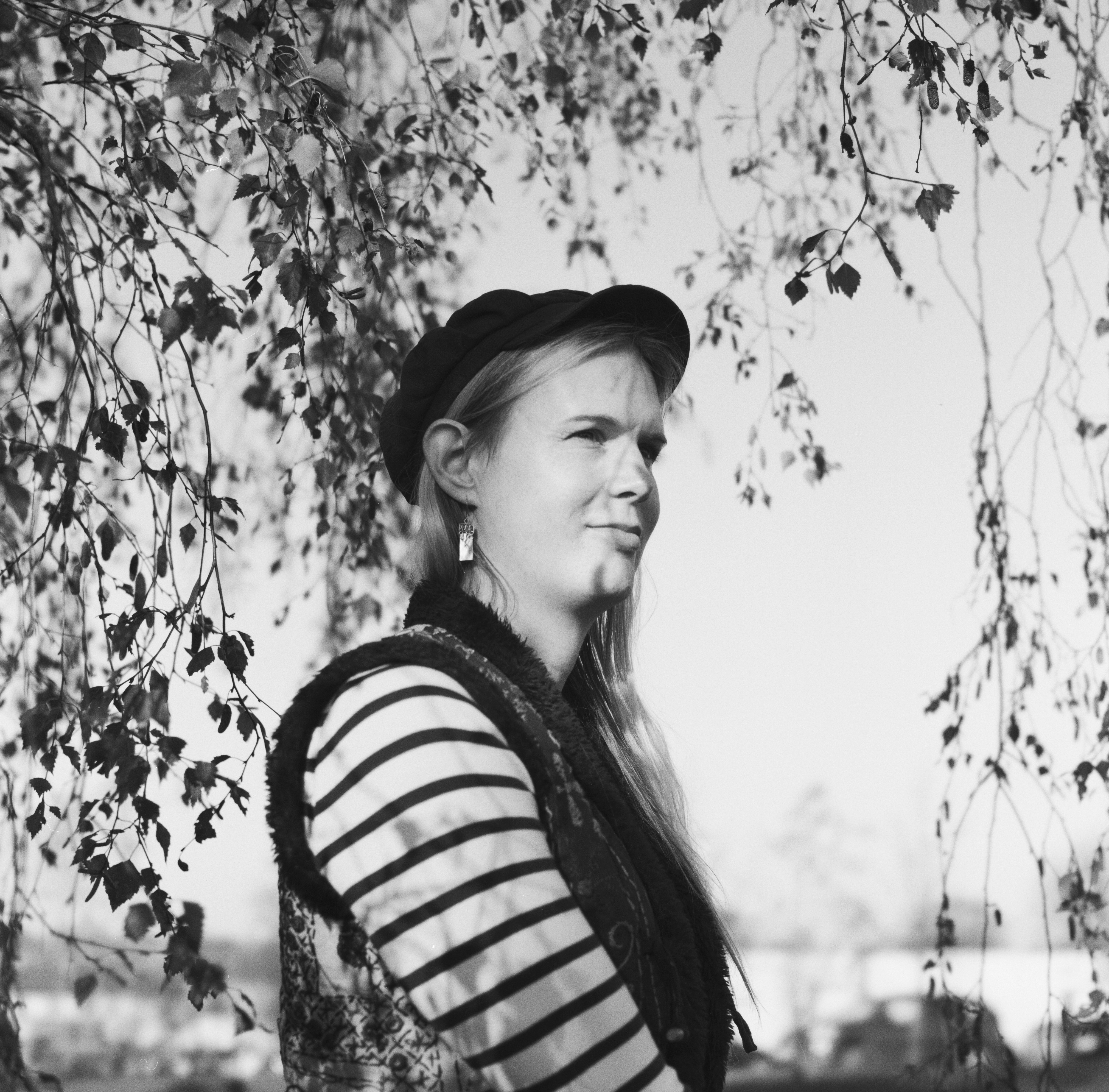 A black and white photograph of a woman from the torse up. She is standing in the midst of branches. She has long blond hair and is wearing a cap.