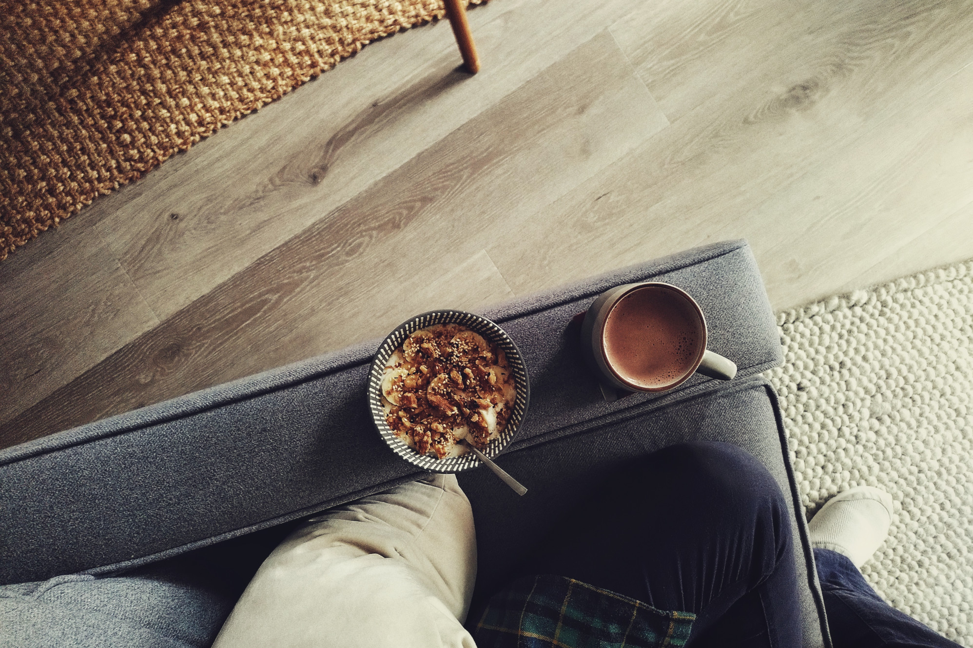 A horizontal colour photograph of a grey sofa armrest with a bowl full of yoghurt, sliced banana, nuts, and spices next to a mug of hot chocolate. On the sofa, a leg in a dark blue trouser is bent. The floor is a light colour with a brown jute carpet at one corner of the image and light grey wooly carpet at another.