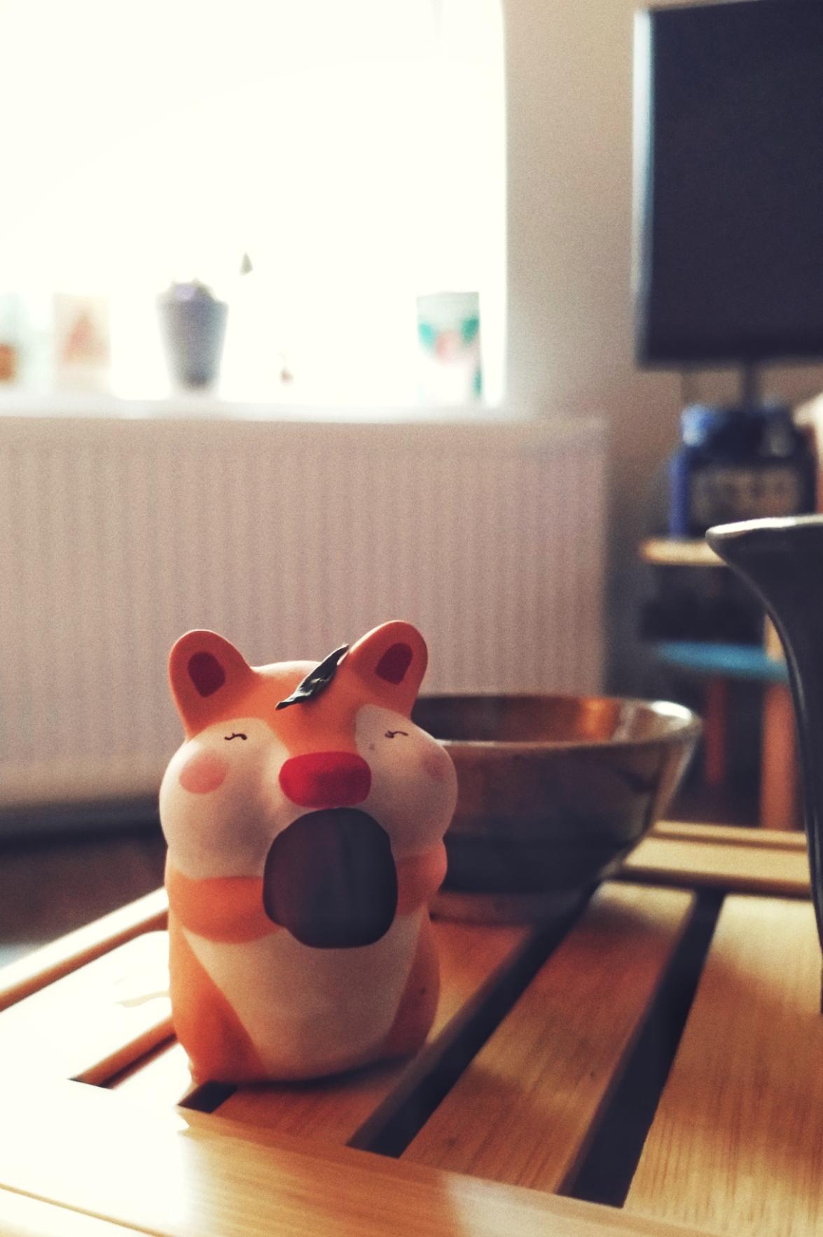 A vertical colour photograph of a hamster tea pet with a mouth full of a big nut. They are on a bamboo tea tray. A green tea leaf rests atop their head. Behind them is a tea cup.