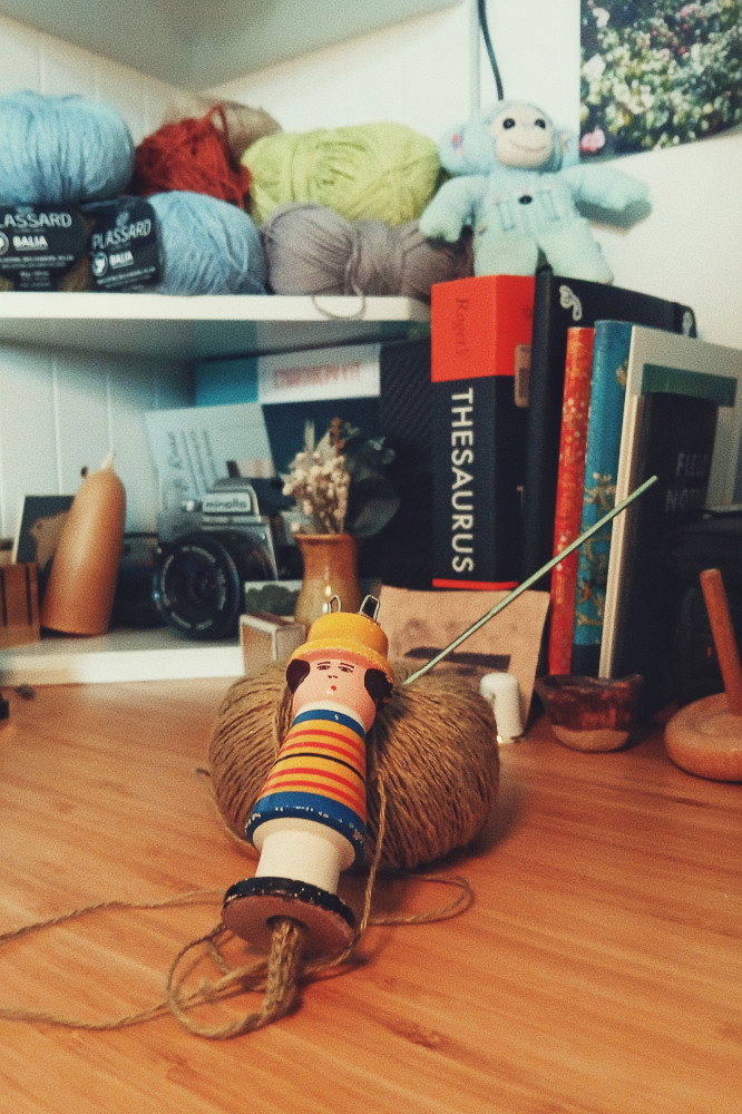 A vertical colour photograph of a wooden figure spool knitting resting on a ball of green yarn. A little bit of the knitted work is coming out at one end. A crochet hooks pokes out of the ball of yarn. Behind is some desk paraphenalia with books, toys, wool, candles, etc.