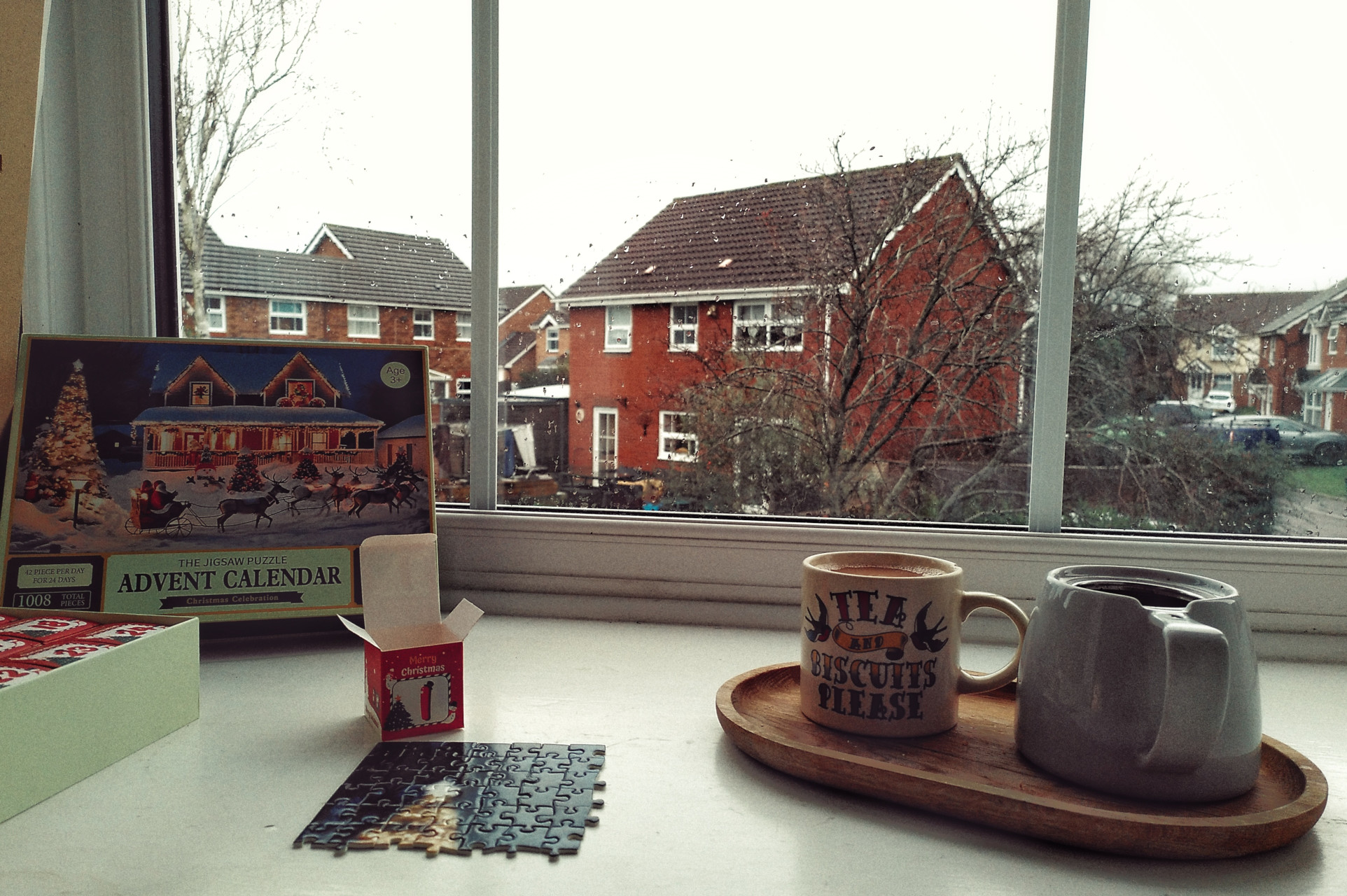 A horizontal colour photograph of a windowsill with the start of a puzzle with a cup of tea next to it. In the background is the puzzle box with the words 'The Jigsaw Puzzle Advent Calendar'. The picture is of father Christmas in the snow pulling in my a house next to a very tall lit tree