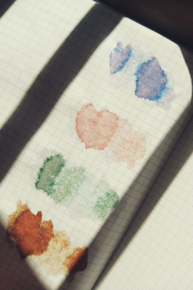 A vertical colour photograph of ink swatches on a notebook with a grid pattern on the paper. This image shows the back of the page with the ink going through the page. Two lines of shadows mark the column of inks. The inks are blue, pink, green, and brown