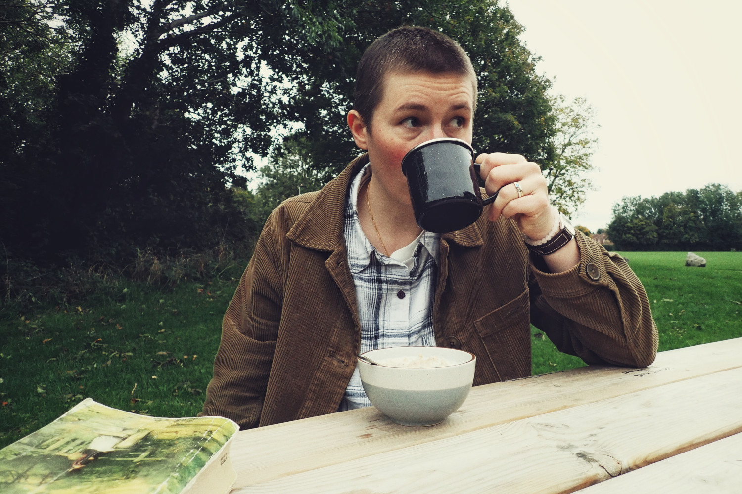 A horizontal colour photograph of me drinking from a black enamel mug. I am a white person with short dark cropped hair. I am wearing a khaki corduroy jacket. My eyes are looking off in the distance. I am in a park.